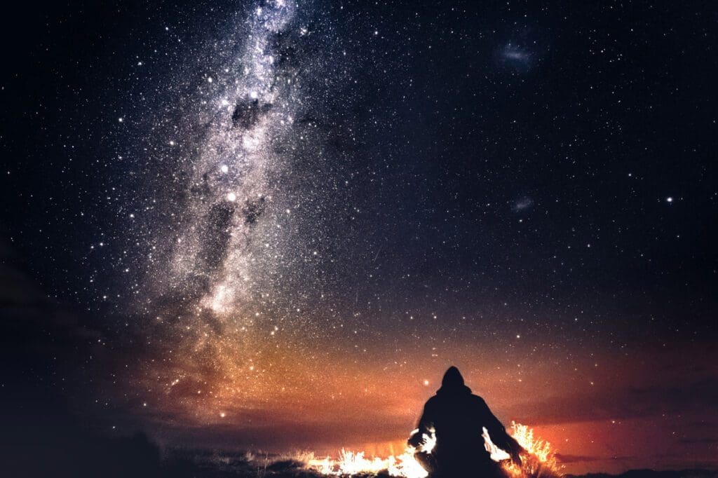 A person sitting in front of a small fire with the milky way showing in the sky