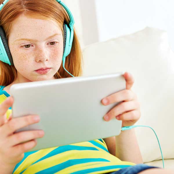 One young child wearing over the ear headphones holds a tablet in her hands and watches the screen.