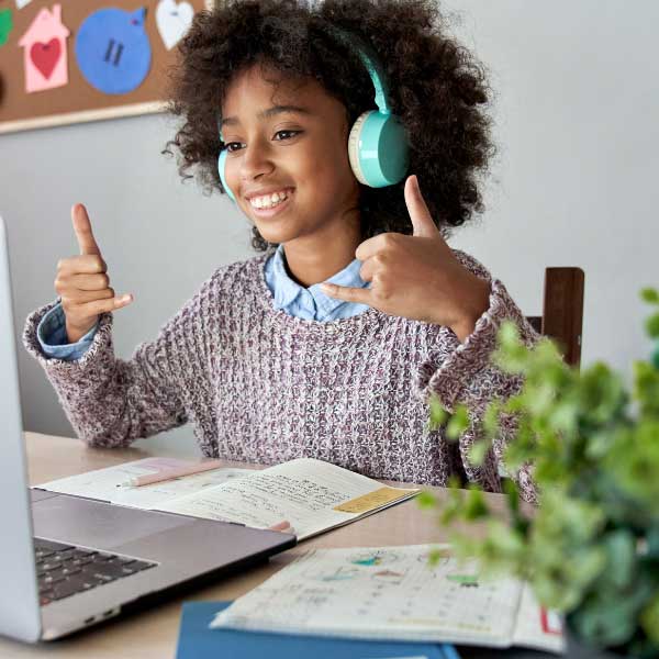A pre-teen girl wears headphones and sits with an open laptop and a book, smiling and giving double thumbs up.