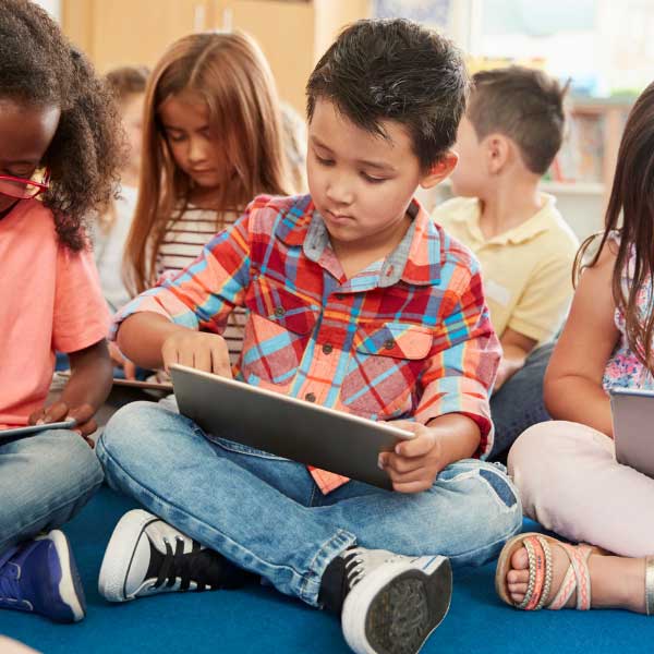 A group of young children sit cross legged on the floor of. a classroom with tablets in their laps.