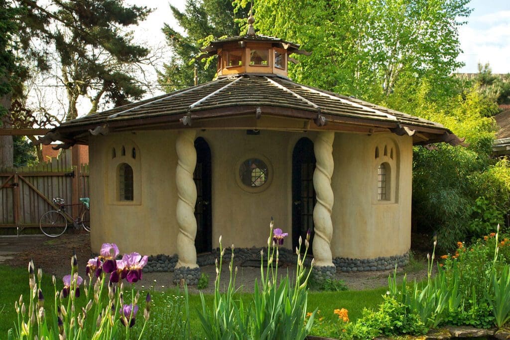 Flowers and trees surround a small yellow clay studio with two pillars in the front. 