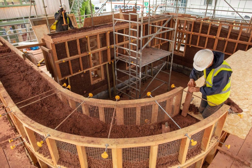 A worker in a white hard hat kneels on a section of plywood as he sets a wall post of a cob structure above the ground.