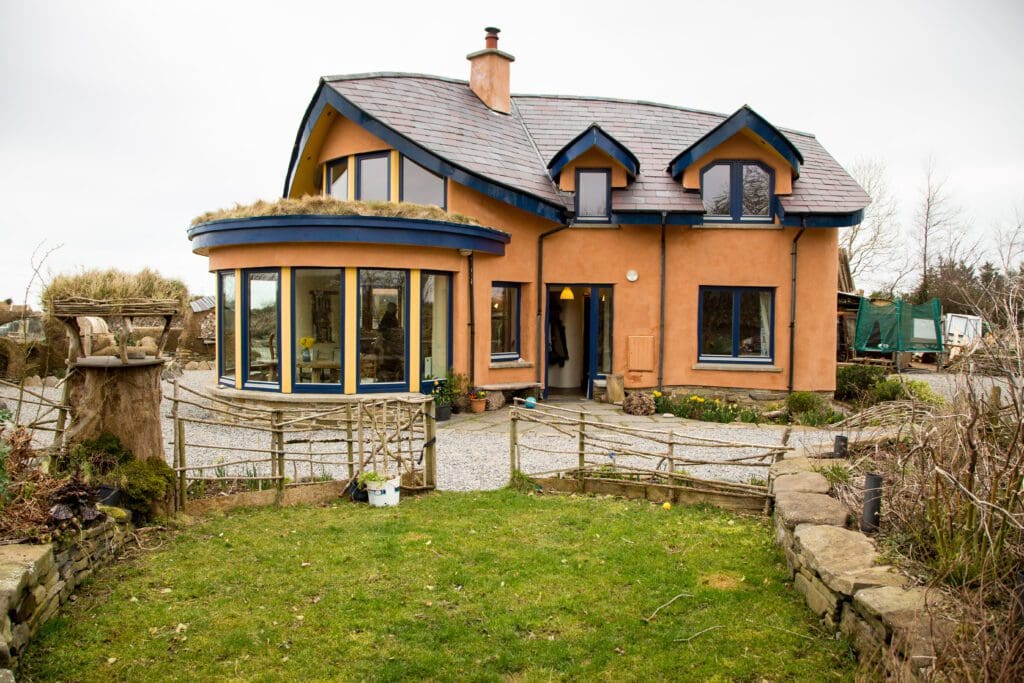 Grass and shrubs surround a 2-story Cob Construction home with a circle of windows under a peaked roof. 