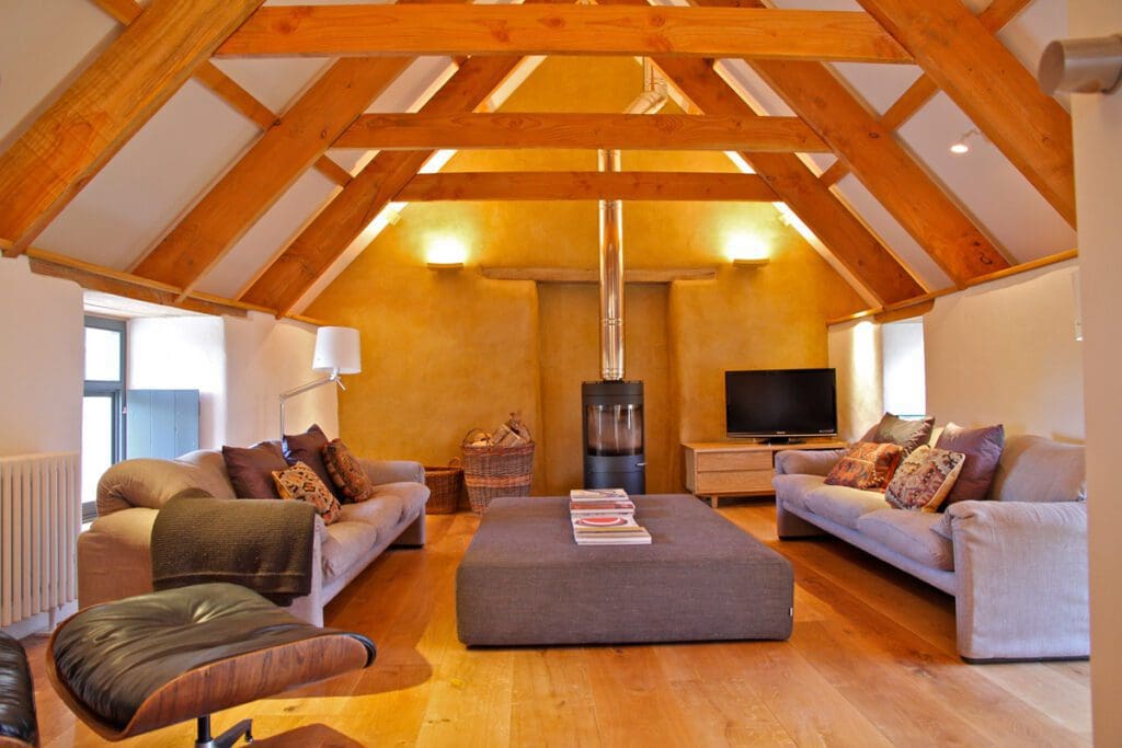 Inside the cozy living room of a cob home, exposed beams sit above 2 couches opposite each other and a wood stove along the north wall. 