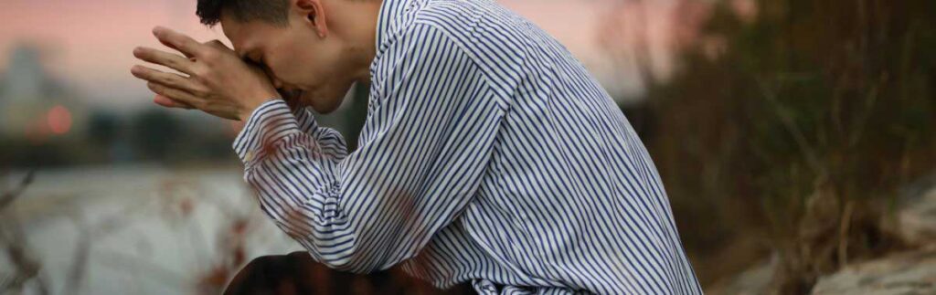 Young man in striped shirt leans on his elbows and puts his head in his hands.