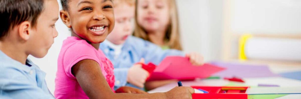 Four young children with art supplies cut paper and draw in a classroom.