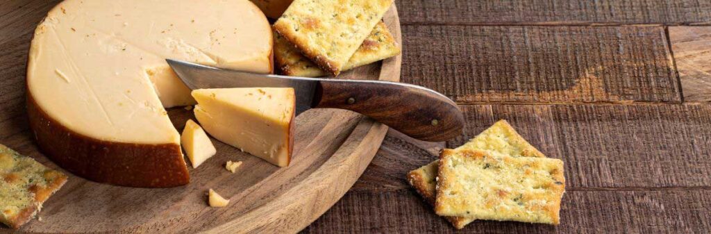 A cheese wheel sits on a round cutting board with a piece cut off with a knife. A few crackers sit on the board and a few more sit on the table.