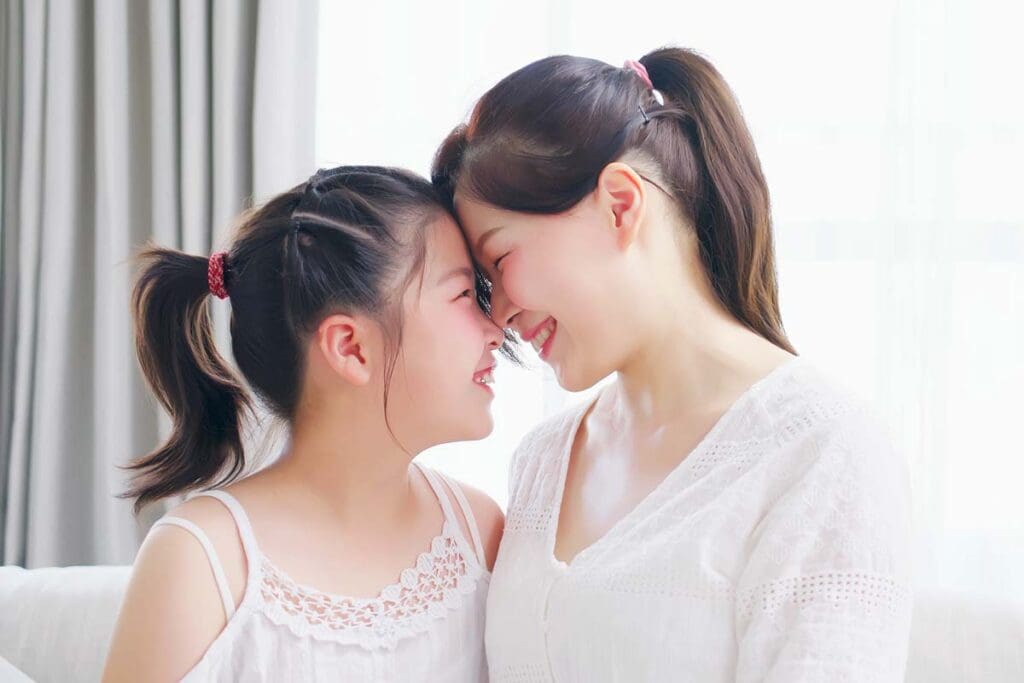 A young woman smiles  with a pre-teen girl, both dressed in white, their foreheads touching.