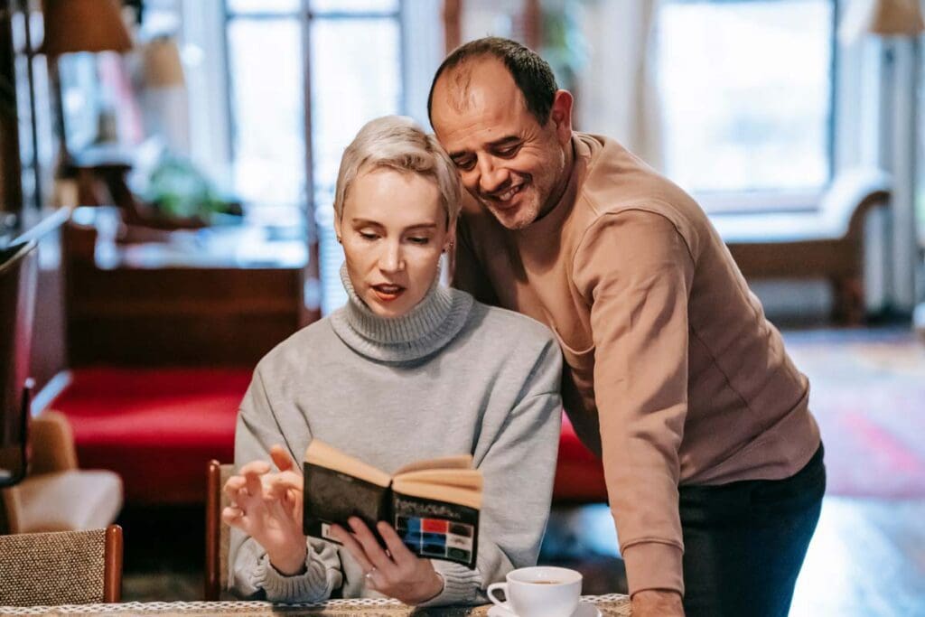 A woman sits at a table with a coffee and reading a novel as a smiling man reads over her shoulder.