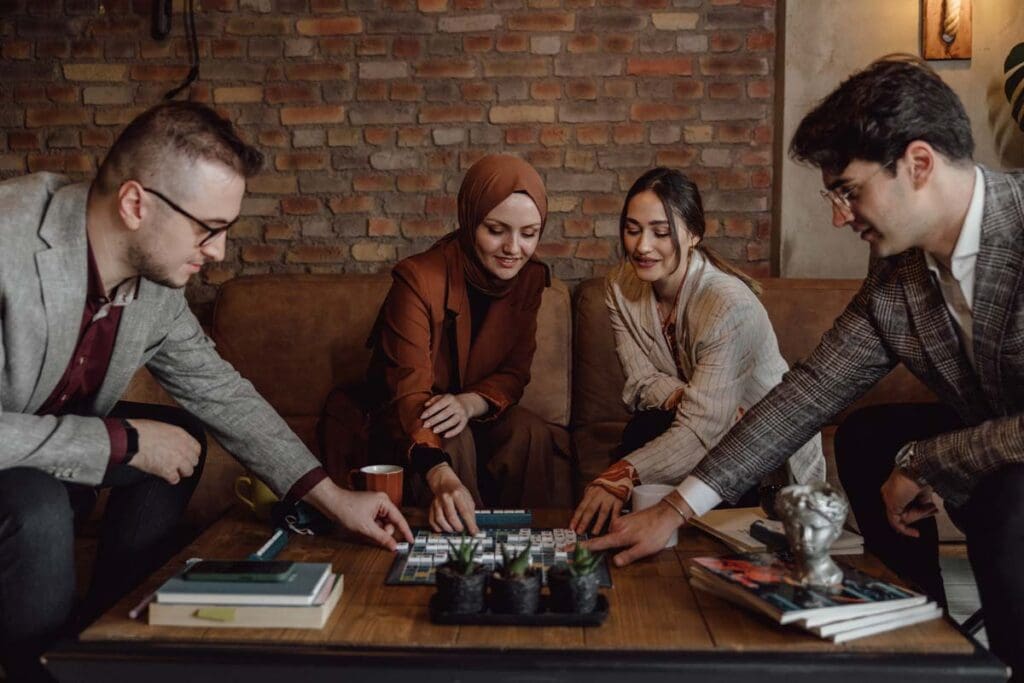 A group of four young, well-dressed, culturally diverse adults sit around a coffee table playing a board game with tiles.