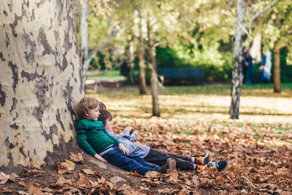 Three small children lay against a large tree on top of a bed of fallen leaves.