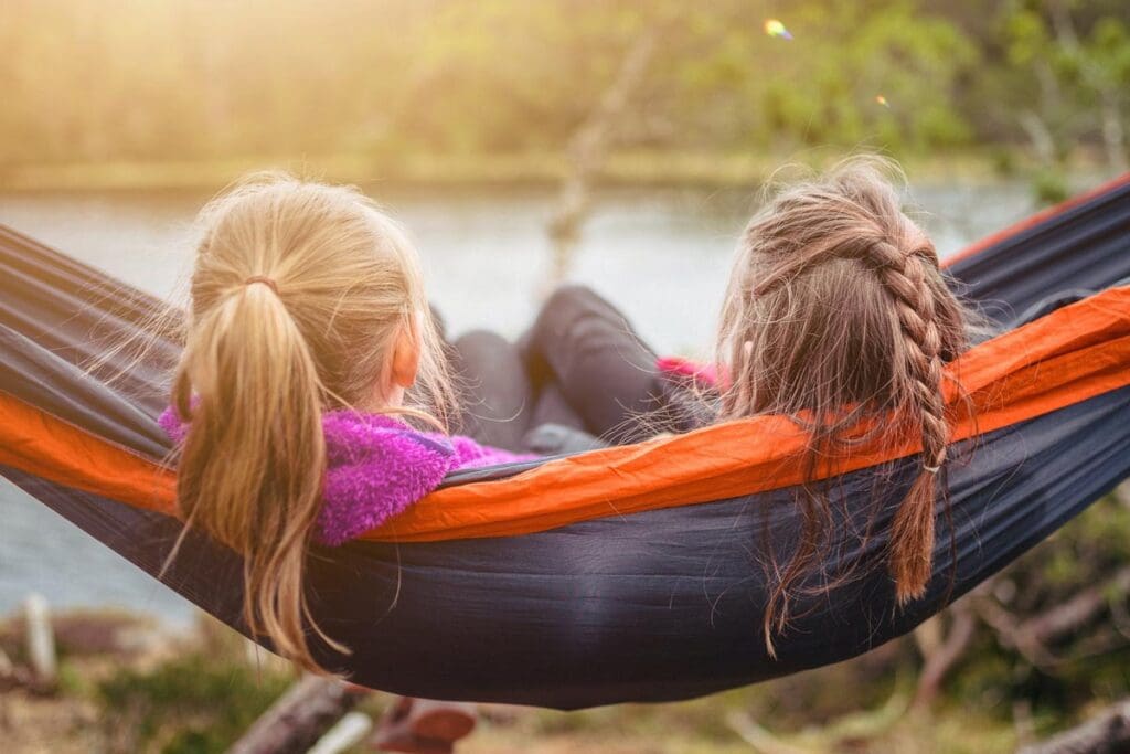 Two girls sit in a hammock look out on a serene lake.
