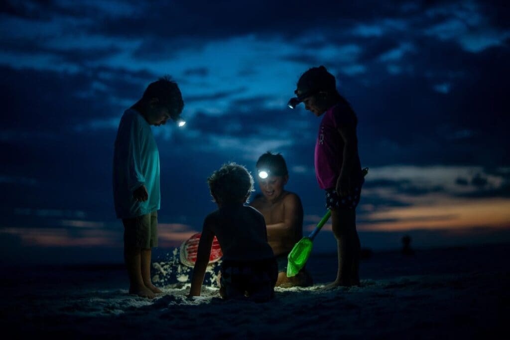 Four small children wearing head lights gather around each other on the beach at night, sifting through the sand