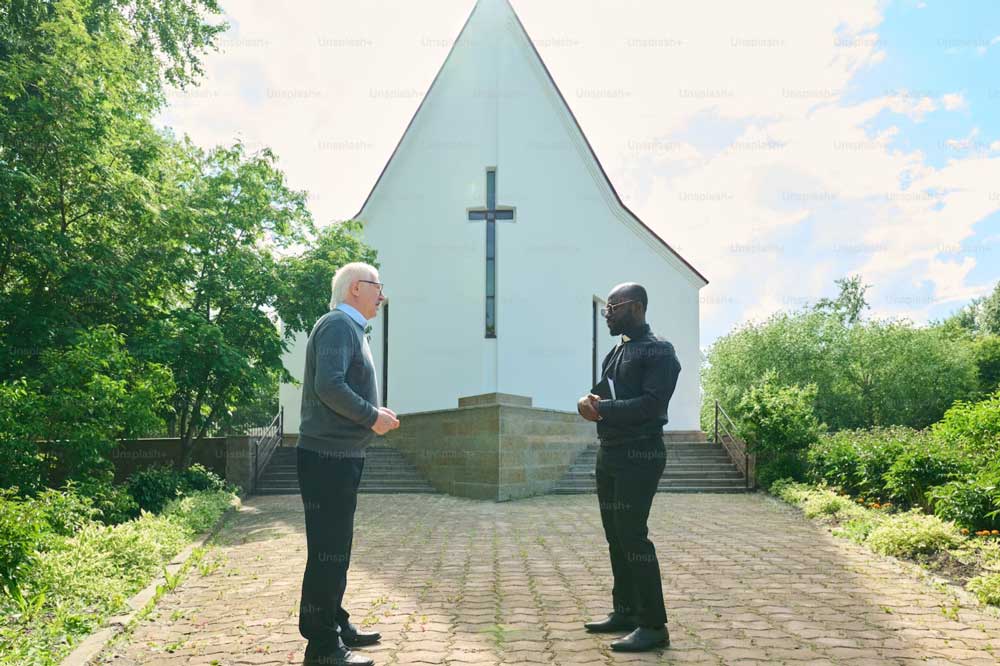 A older caucasian man and an African American man stand across from each other on the walkway in front of a baptist church.