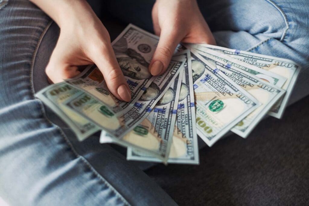 A person is sitting cross-legged, holding a selection of $100 US bills fanned out.