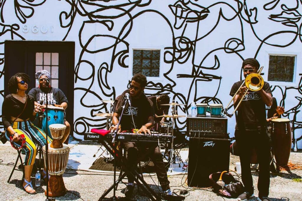 A band (drums, keyboard and sax) perfomrs outside a building decorated by art.