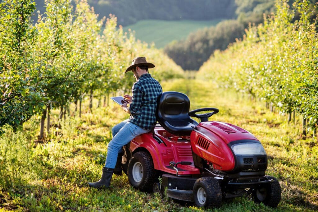 A farmer in a blue plaid shirt, brown hat and jeans holds an iPad as he relaxes on the side of a red riding lawnmower in the field.
