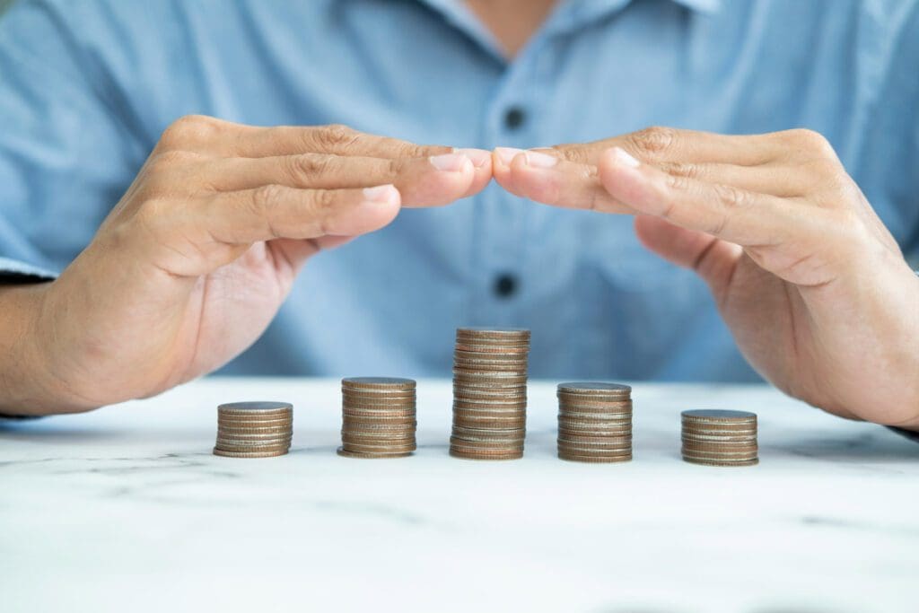Five stacks of coins are on a table under a man in a blue shirt with his fingertips touching.