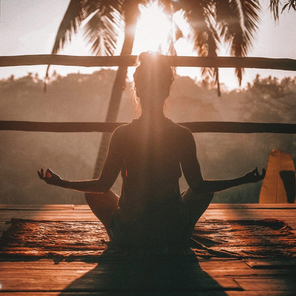 A young woman sits on a deck in a yoga pose on the floor, basking in the early morning light.