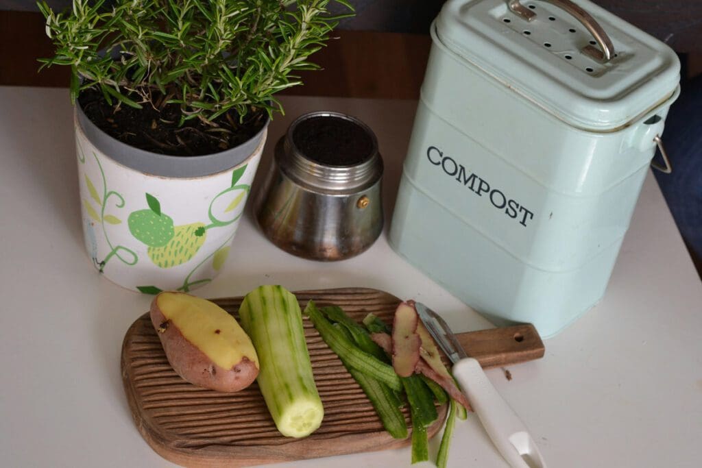 Compost bin and plants in planters with soil sit on a countertop with a half-peeled potato and cucumber. The skins are sitting beside them on a cutting board.