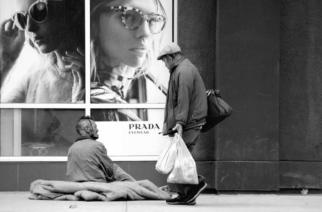 A black-and-white photo shows a man sitting on a blanket outside a Prada store that features ads of women wearing sunglasses.
