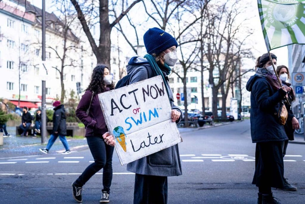 Masked activists demonstrate on a city street; one holds an Act Now or Swim Later sign.