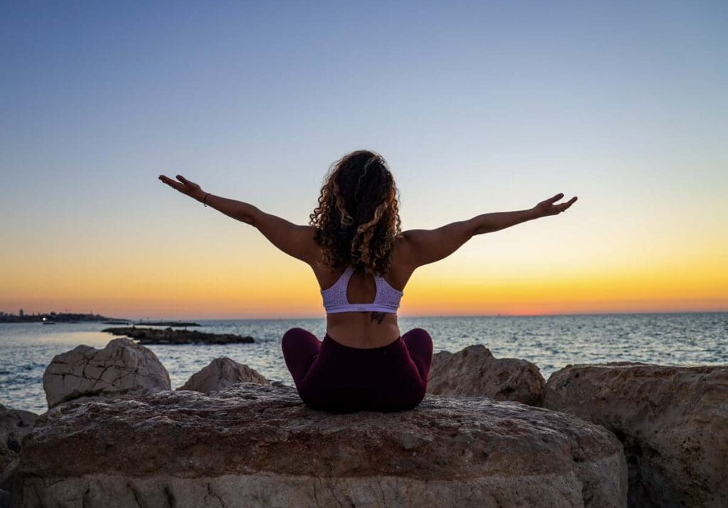 A young woman sits on a rock outcropping doing yoga in front of the ocean at sunrise.