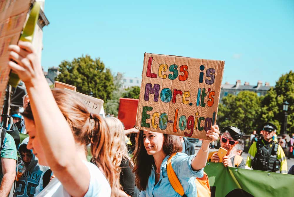 A group of activists protest outside; one woman holds a colorful Less Is More It's Eco-Logical sign.