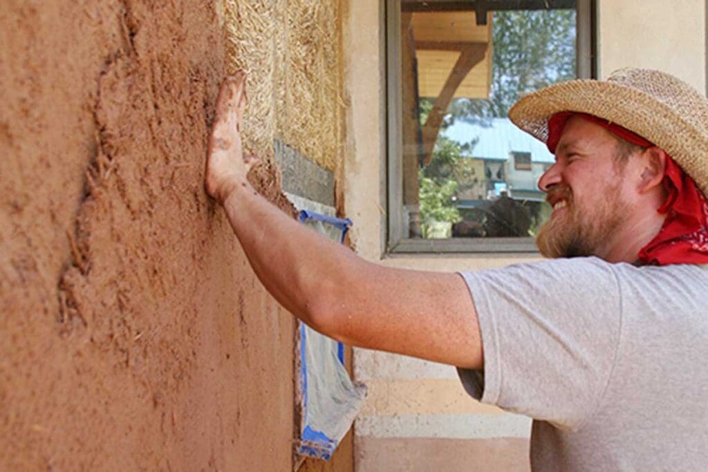 A gentleman in a straw hat applies straw and plaster to the inside walls of a home.