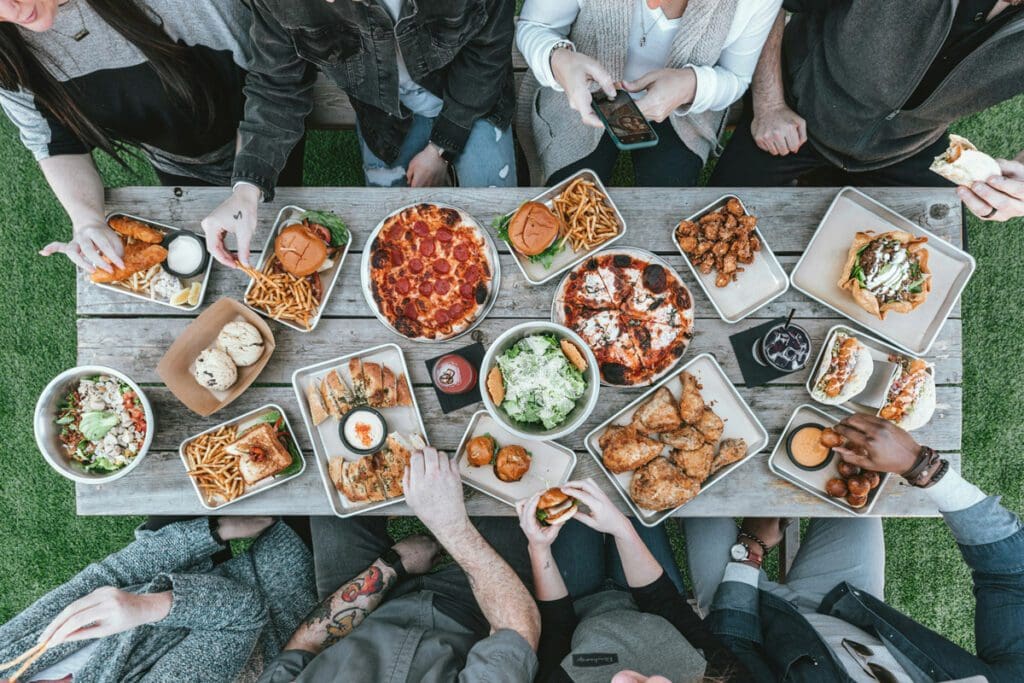 Picnic table full of meals of pizza, hamburgers and fries, chicken and salads with four eaters.