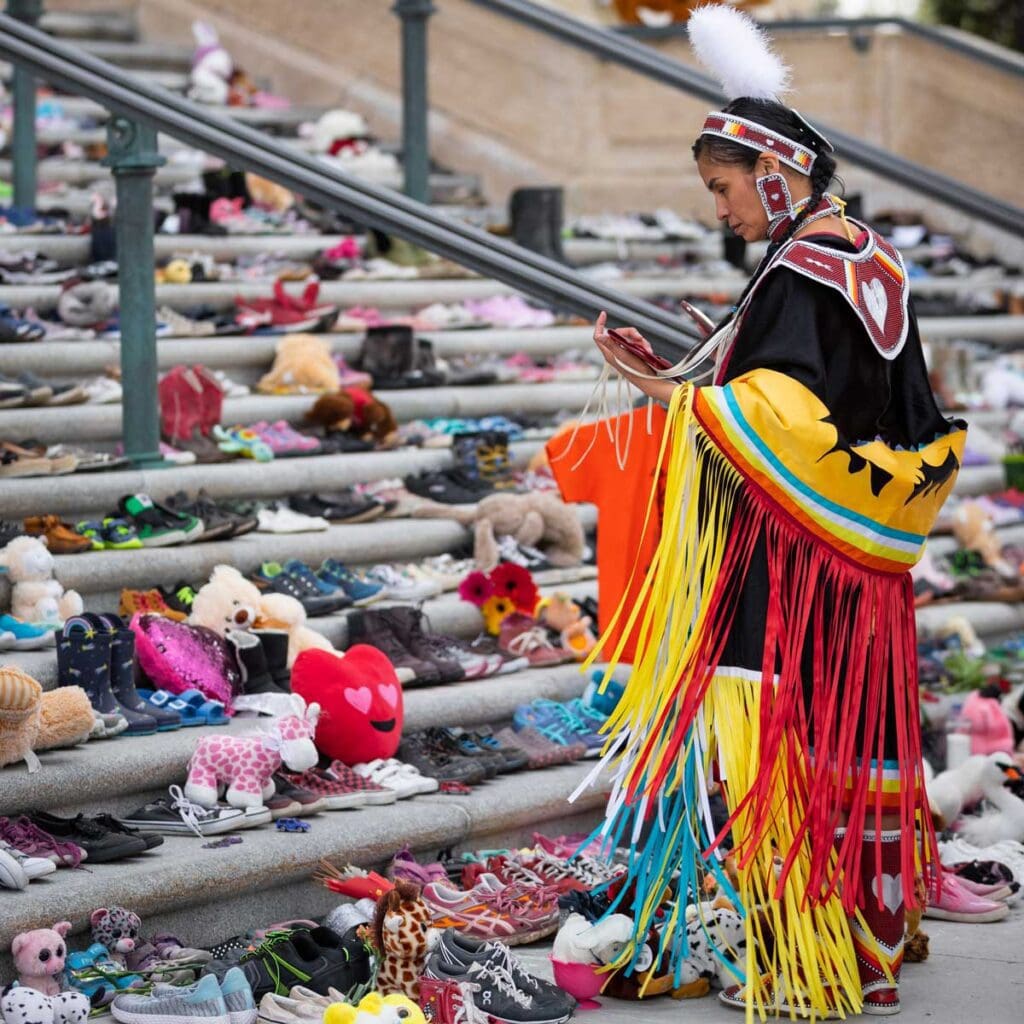 Indigenous woman in cultured garb pauses in front of the steps of a building with rows of flowers, stuffed animals and footwear.