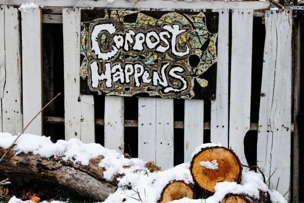 A Compost Happens sign sits on an older white wooden fence in the snow.