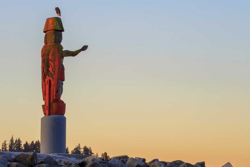 An indigenous carving on a concrete base sits on rocks with its arm outstretched.
