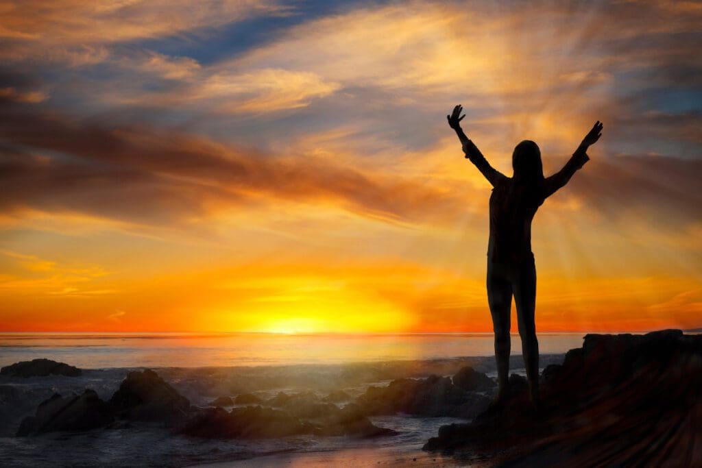 Silhouette of a person overlooking the ocean and the sunset, while standing on rocks by the beach