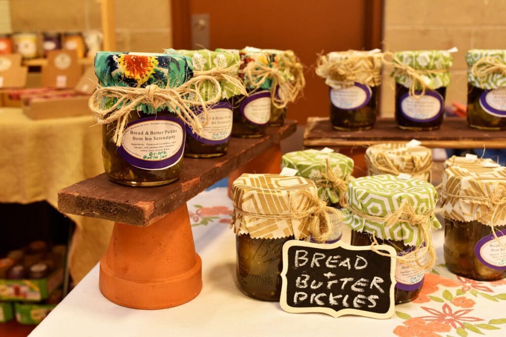 A display table with preserves (Bread & Butter Pickles) with assorted linen lid coverings decorated with twine. A sign on the table reading "Bread & Butter Pickles."