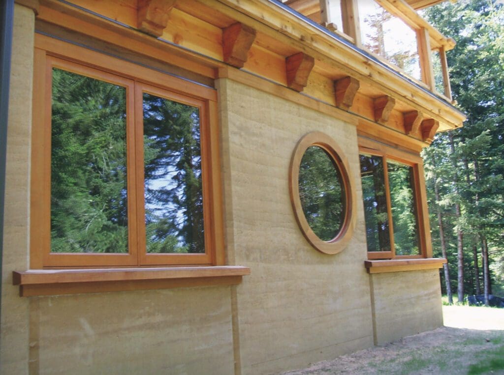 The image showcases a beige-colored building featuring wooden-framed square and circular windows, set against the lush backdrop of green trees.