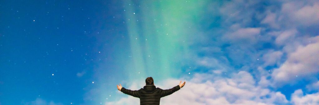 A person stands with arms outstretched under a night sky illuminated by the Northern Lights, featuring shades of green and blue against a backdrop of stars and clouds. The individual appears to be wearing a dark coat and is facing the sky, evoking a sense of awe and wonder suitable for the scene described in Social_Excerpt_Introduction-to-Power.