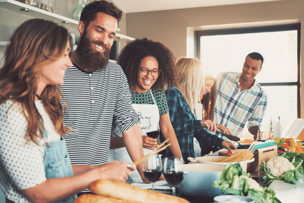 Six people gather around a kitchen island, smiling and prepping a past dinner while drinking wine.