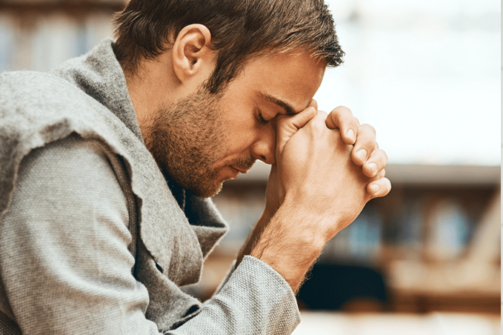 A man leans with his clasped against his forehead; his eyes closed and his head bowed.