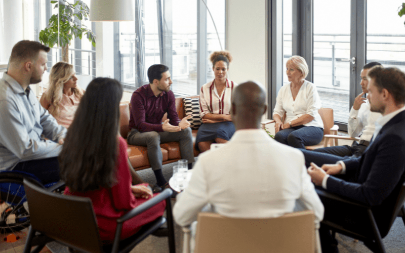 A diverse group of well-dressed people sit on a couch and chairs in a circle collaborating