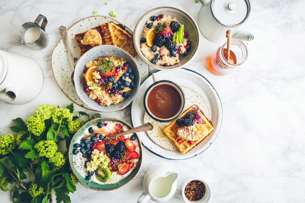 Breakfast includes waffles, oatmeal, fruit, nuts, honey, tea, and milk on the table.