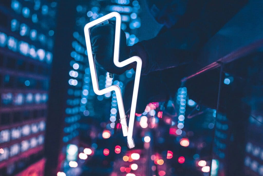 In an urban night setting, a hand holds up a glowing white neon lightning bolt sign. The background is filled with blurry city lights and buildings, highlighting the vibrant and energetic atmosphere.