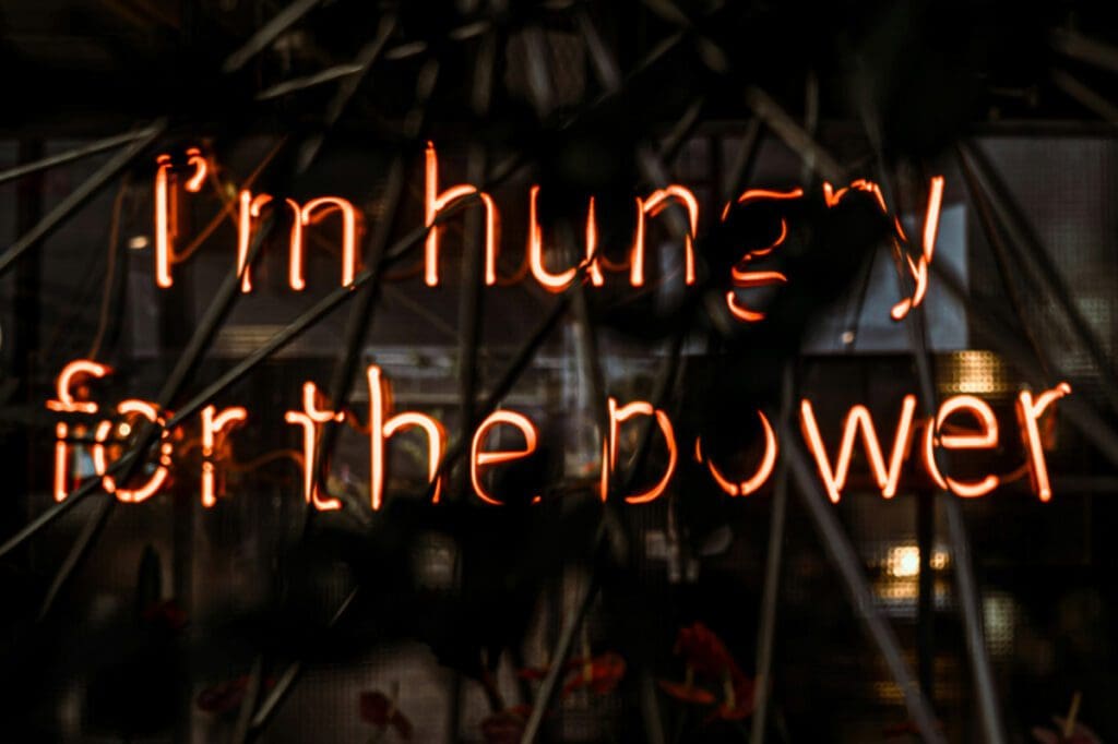 The image titled "raphael-renter-raphi_rawr-frCuMW5TD58-unsplash" features a glowing neon sign displaying the text "I'm hungry for the power," set against a dark background with blurred rods and reflections in the foreground.