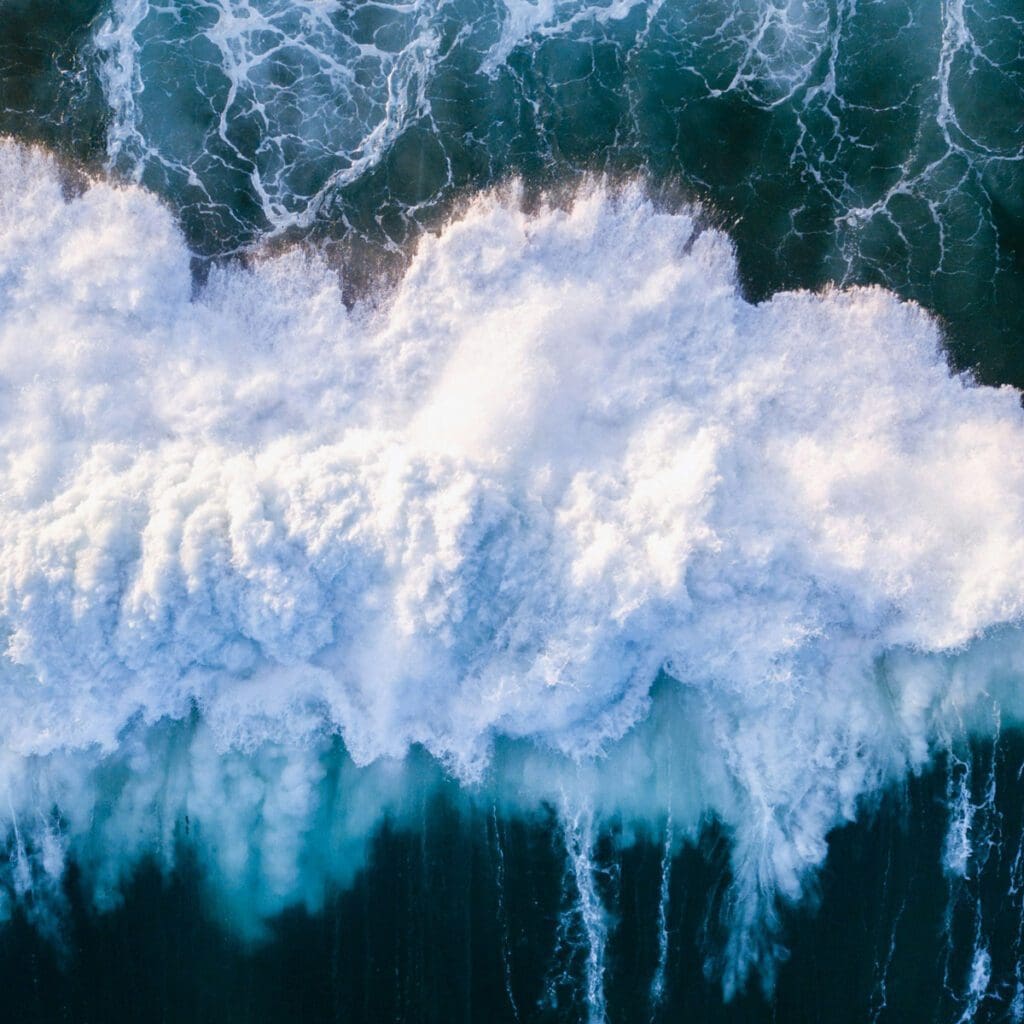 Aerial view of ocean waves crashing against each other, creating a frothy white foam. The deep blue water contrasts with the vibrant foamy crests, showcasing the dynamic and powerful movement of the sea.