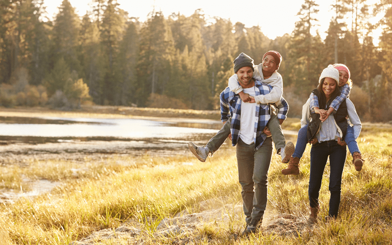 A mother and father give their children piggy back rides down a path next to a lake.
