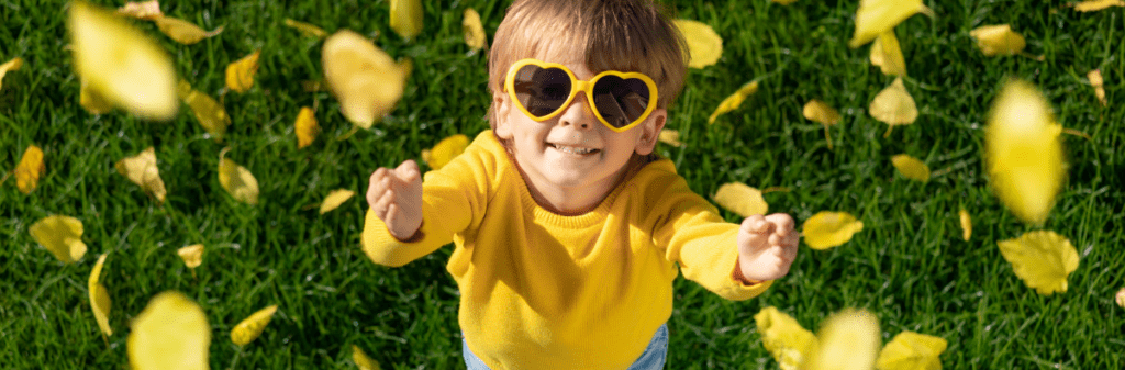 Young boy in yellow heart sunglasses reaches for leaves falling from overhead.