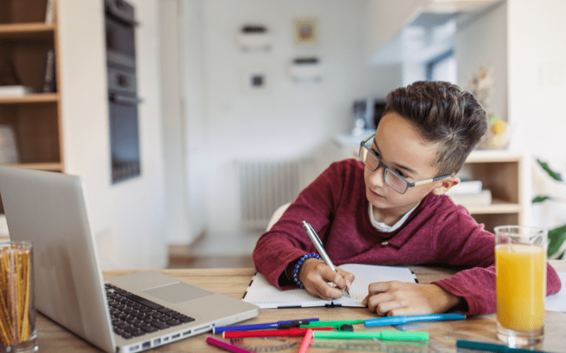 A young boy in glasses sits at a table in front of a laptop writing on a piece of paper (doing homework).
