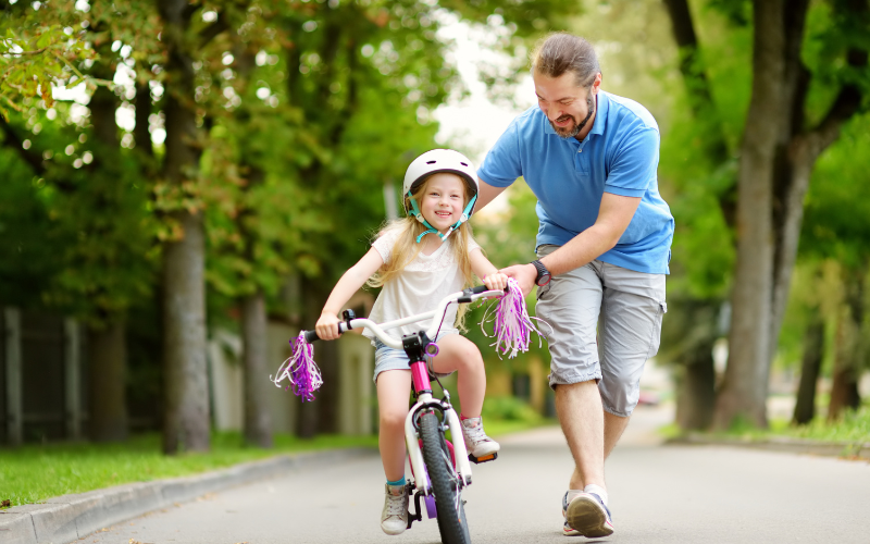 A young father pushes his daughter down the street on a bike.