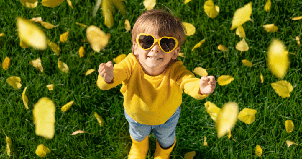 Young boy in yellow heart sunglasses reaches for leaves falling from overhead