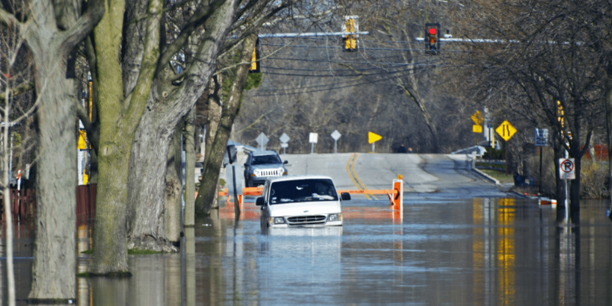 A white truck is partially submerged on a flooded street with trees lining the road and traffic lights in the background.